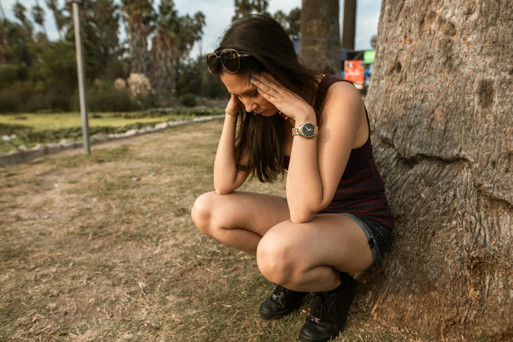 Dizer “não” sem culpa: o limite que acalma a ansiedade A woman kneels by a tree holding her head, expressing stress or anxiety in an outdoor park.