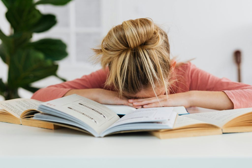 A young girl falls asleep on a desk surrounded by open books, illustrating study fatigue.